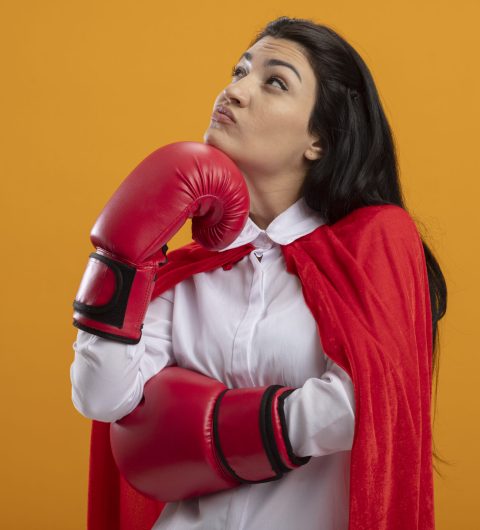 thoughtful young caucasian superhero girl wearing box gloves putting hand on chin looking up isolated on orange background with copy space